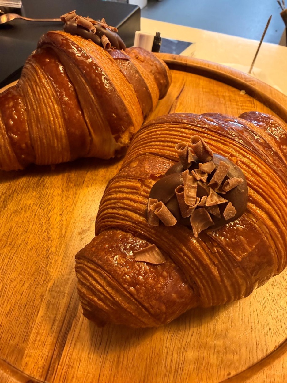 Two glossy chocolate croissants topped with chocolate cream and curls on a wooden board