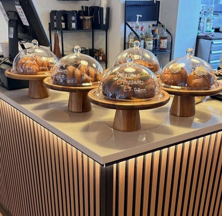 Croissants under glass domes on wooden cake stands on the Thrive counter, including pistachio and custard croissants