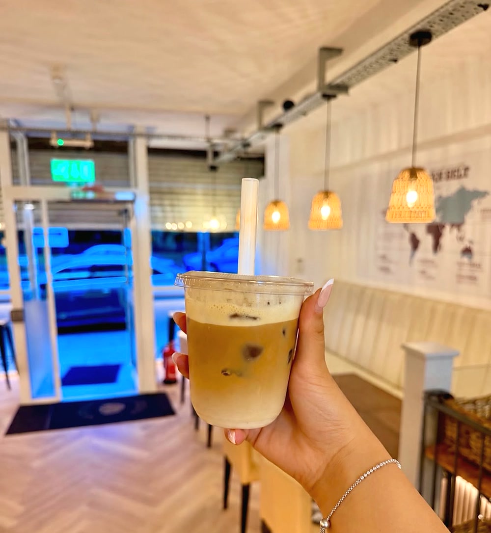 An iced latte held up inside Thrive coffee shop, with rattan pendant lights and booth seating